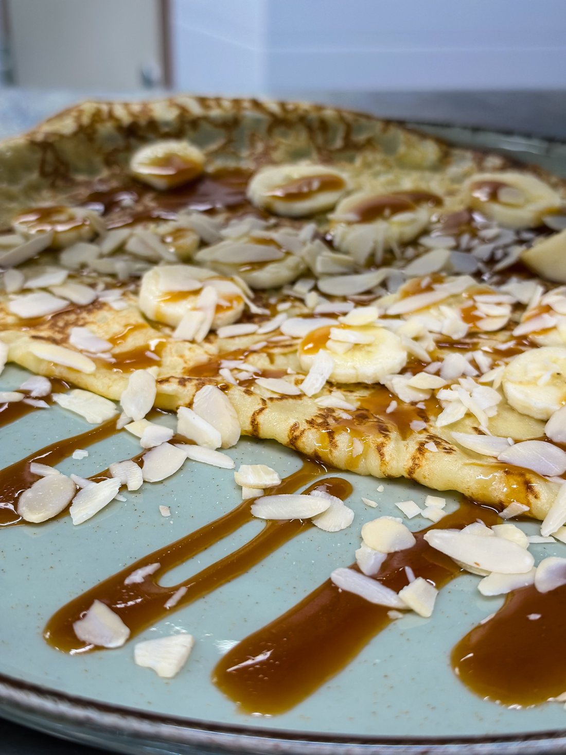 Une crêpe de froment garnie de rondelles de banane et d'amandes effilées, nappée de caramel au beurre salé, sur une assiette vert d'eau