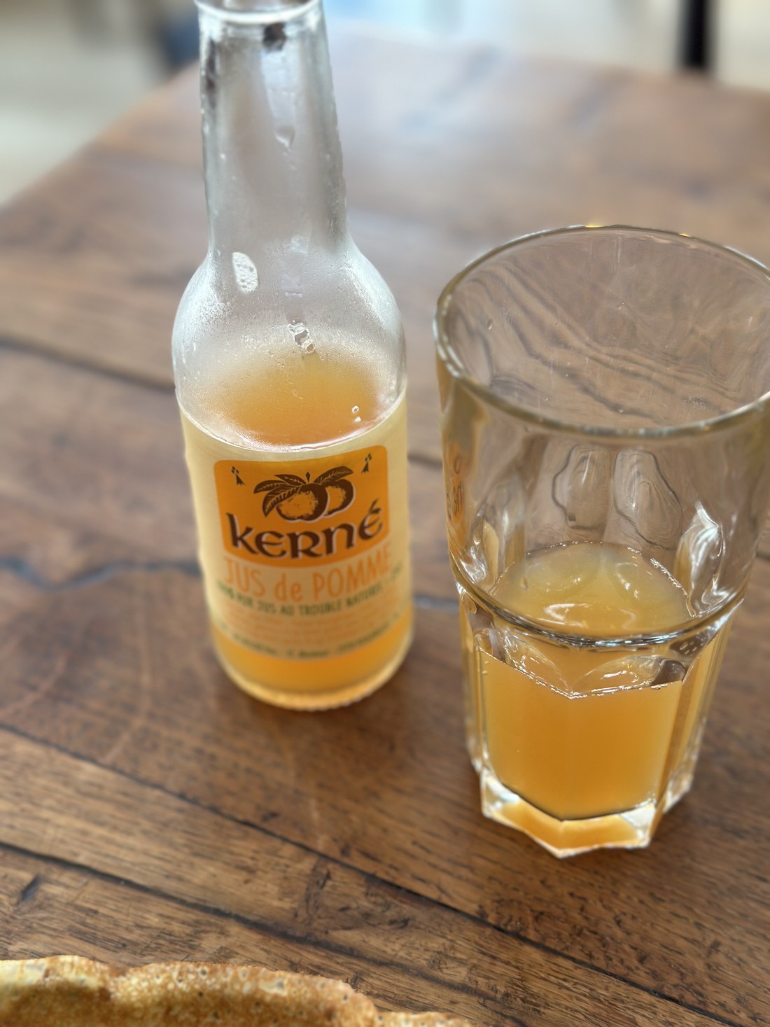Une bouteille de jus de pomme artisanal Kerné et un verre servi, posés sur une table en bois à côté d'une galette